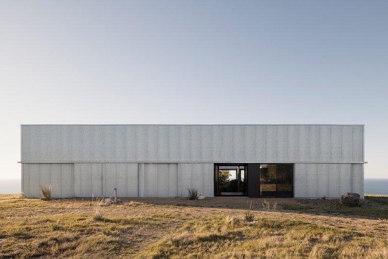 Long horizontal volume clad in vertical corrugated metal with central recessed entry at dusk