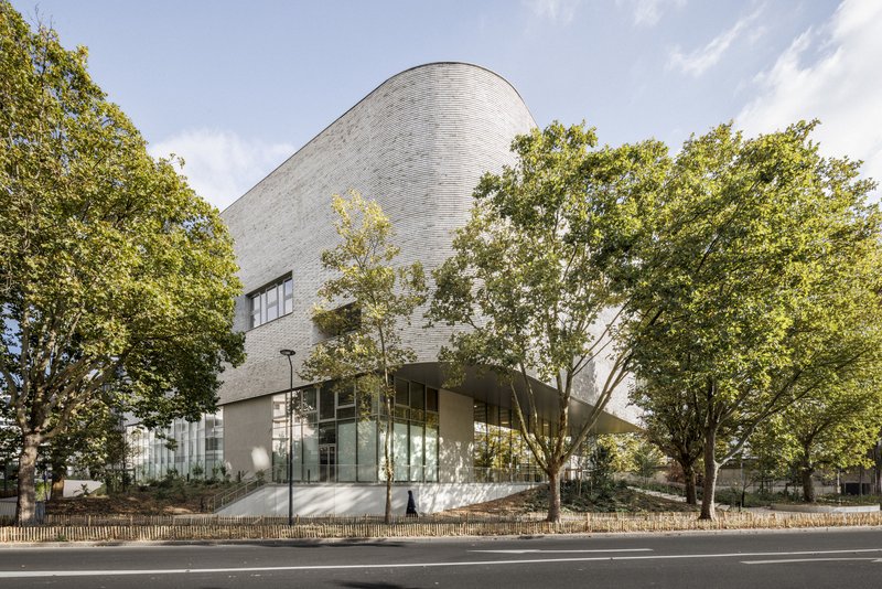 Street view of the curved brick volume rising above a glazed ground floor framed by trees