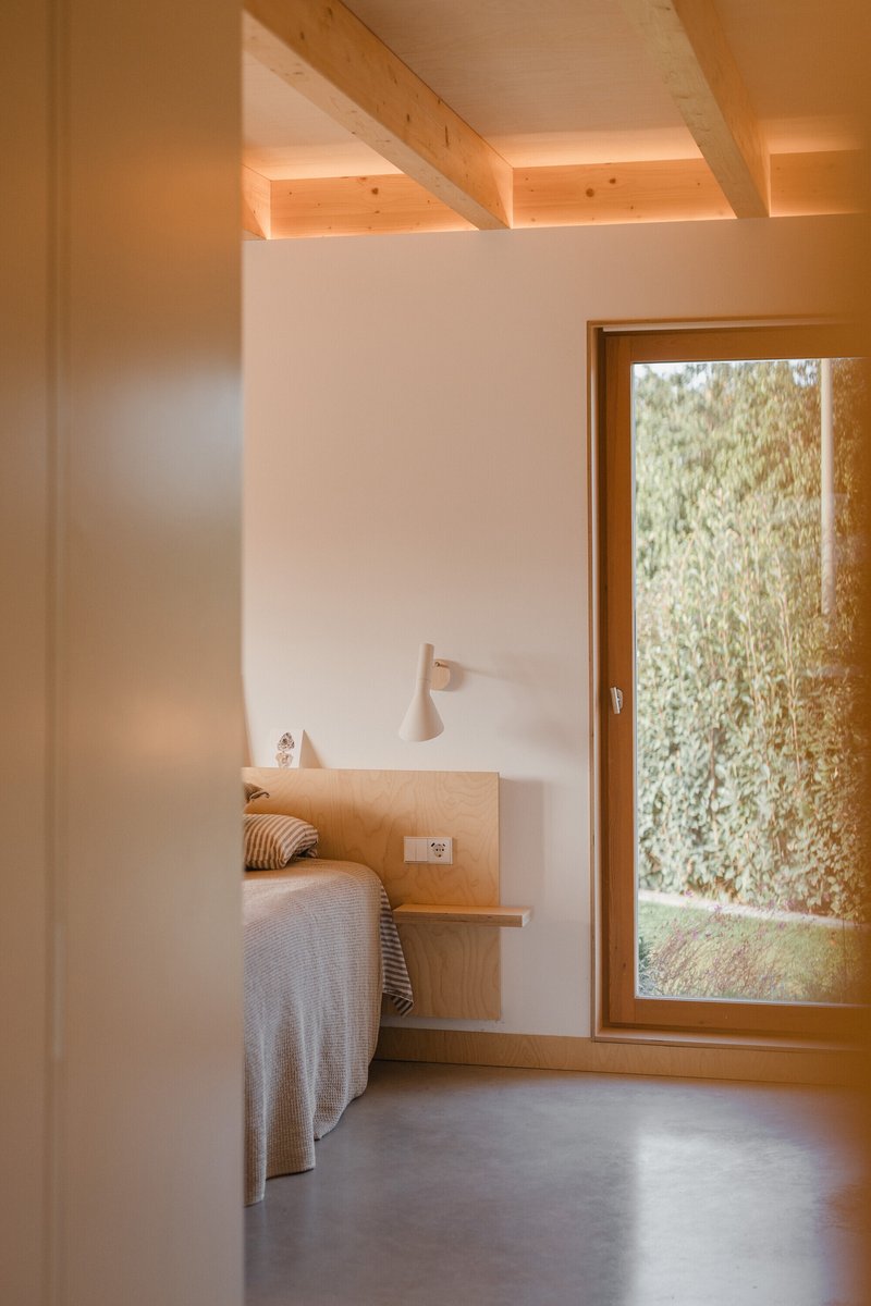 Bedroom with plywood bed frame and timber-beamed ceiling lit by afternoon sunlight through a garden-facing window
