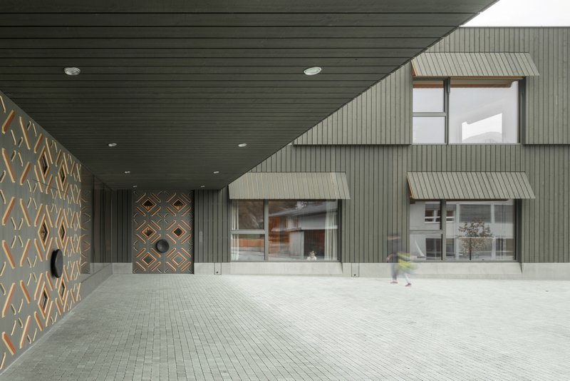 Covered entrance courtyard with timber-paneled ceiling and patterned wall adjacent to metal-clad facade volumes