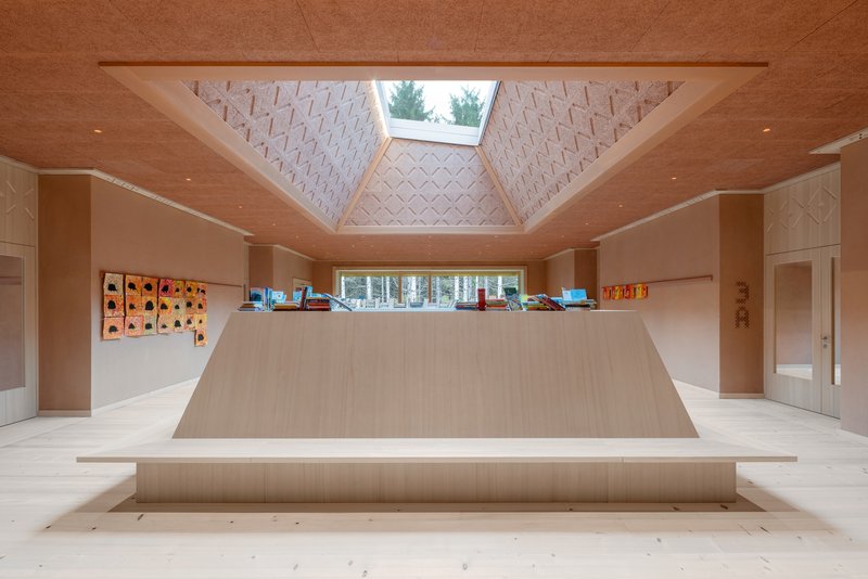 Light timber interior hall with pyramidal skylight and geometric acoustic ceiling panels above a raised platform