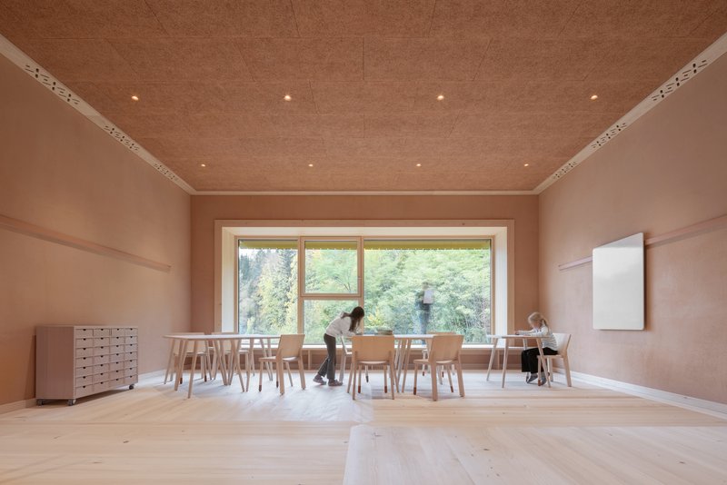 Timber-lined classroom with horizontal window framing hillside vegetation and children seated at low tables