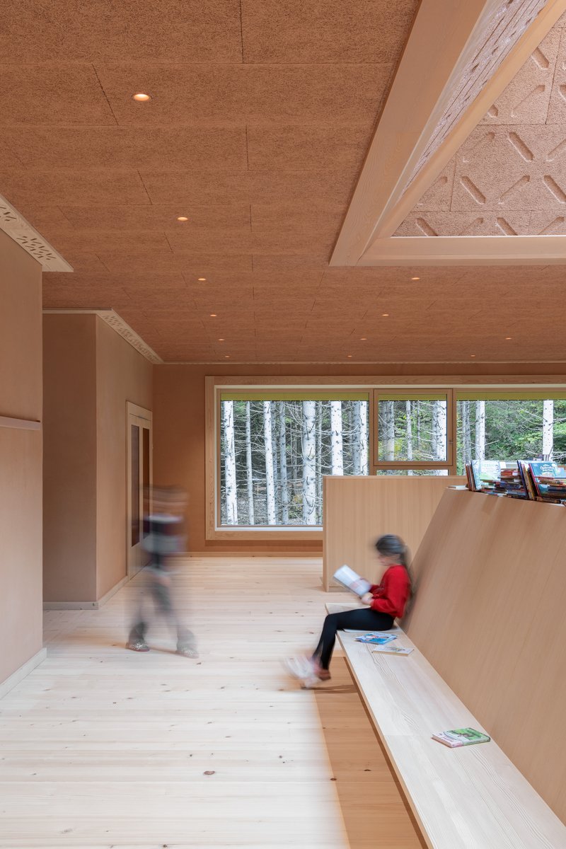 Interior corridor with child in red jacket seated on timber bench near window overlooking birch forest