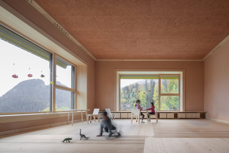 Light-filled classroom with terracotta ceiling panels, pale wood floors, and children playing near large windows overlooking forested hills