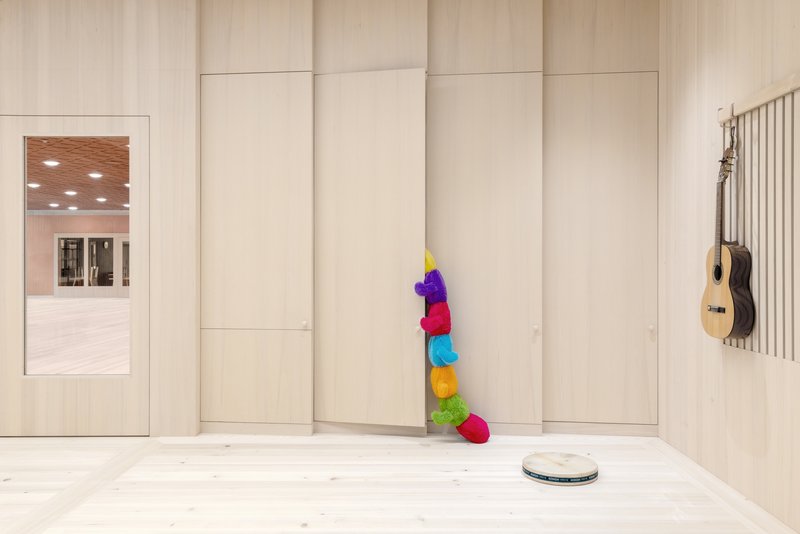 Corridor wall with recessed panels, a guitar mounted on vertical slats, and colorful caterpillar toy against pale wood surfaces