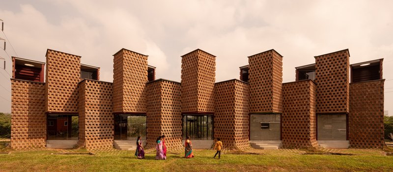 Perforated brick facade with cylindrical columns and glass openings as people walk across the lawn