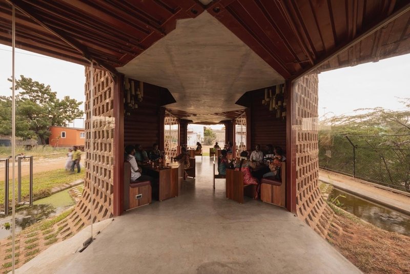 Covered passage with timber ceiling panels and concrete beams between perforated brick walls with seated visitors