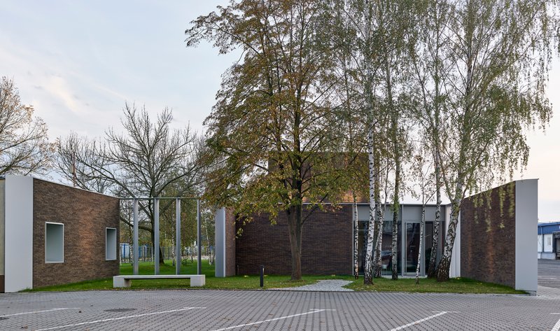 Street view of the dark brick and white panel volumes with birch trees and benches in a courtyard setting
