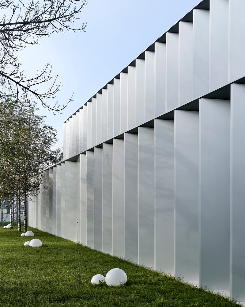 Translucent vertical panel facade with triangular shadow reveals beside a sloping lawn and bare trees