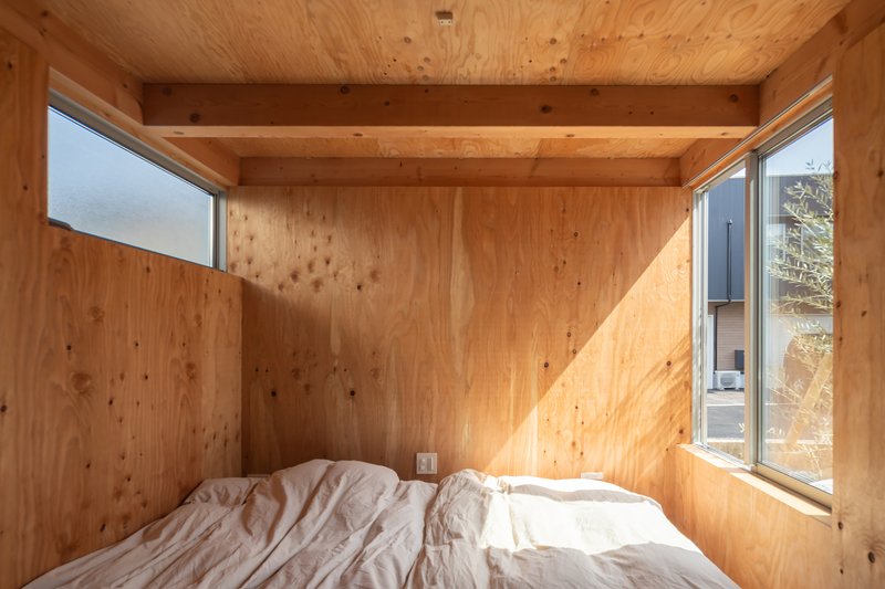 Plywood-lined bedroom with exposed beams and corner windows casting sunlight onto a white futon