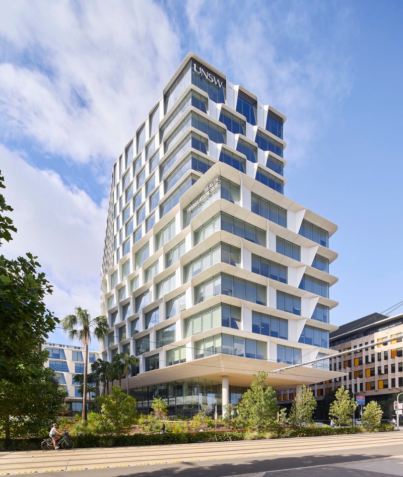 Street-level view of the cantilevered white facade with cyclist passing palm trees and planted beds