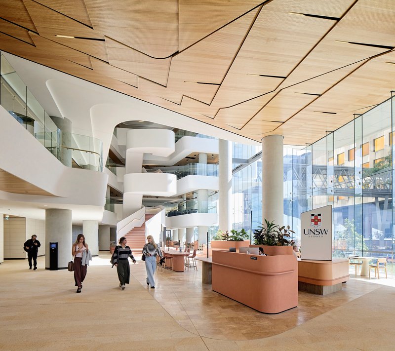 Atrium with terrazzo flooring, white curved balconies, timber ceiling planes and students entering beneath glazed facades