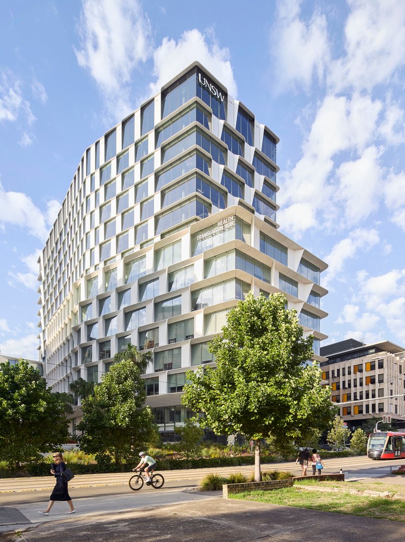 Rectilinear tower with offset windows and white frames rising above street-level trees and a cyclist passing