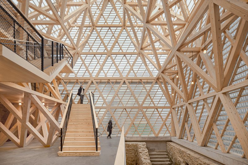 Interior atrium with diagonal timber lattice structure and wooden staircase above excavated stone foundations