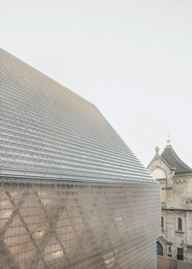 Faceted glass tile roof volume adjacent to a historic stone church tower under grey skies
