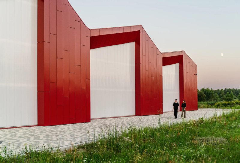 Red metal-clad gabled volumes with recessed white entries and two people walking at dusk