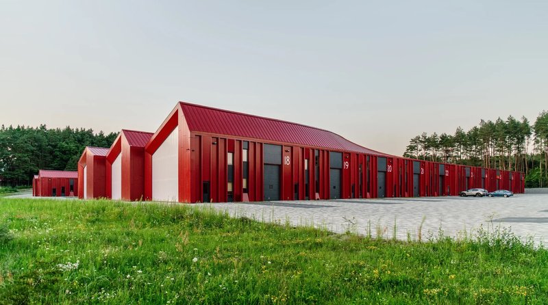 Long facade of red metal cladding with vertical window slots and a pine forest beyond