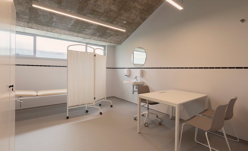 Clinical room with white-tiled walls, exposed concrete ceiling and a desk area by the window