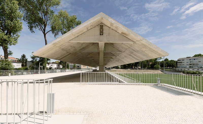 Cantilevered concrete roof with exposed formwork hovering above white railings and a football pitch