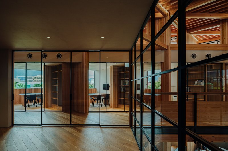 Interior hallway with black-framed glass partitions revealing adjacent timber-clad rooms and exposed joists in evening light