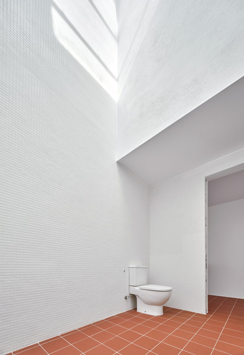 White-tiled bathroom with clerestory window casting angular shadows above terracotta floor and toilet fixture