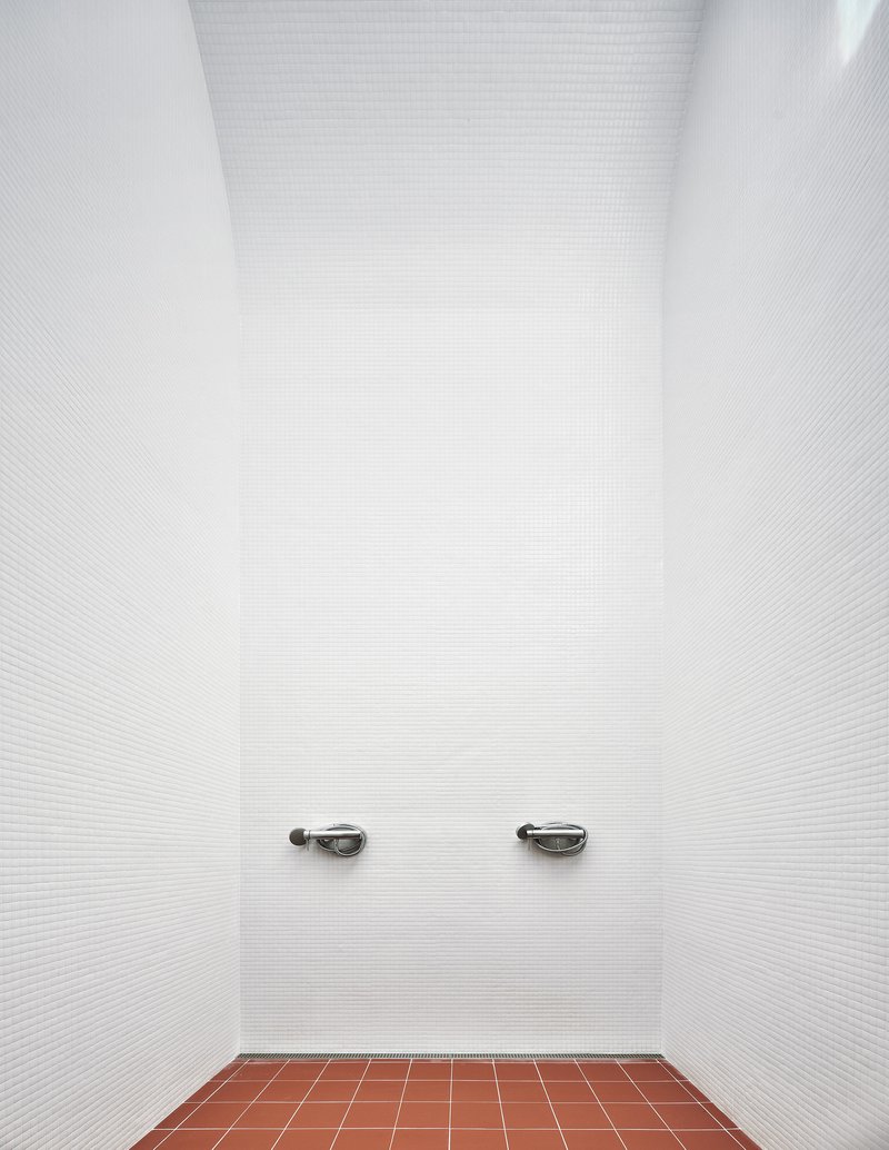 Narrow shower alcove with white mosaic tile walls and two chrome faucets above terracotta floor