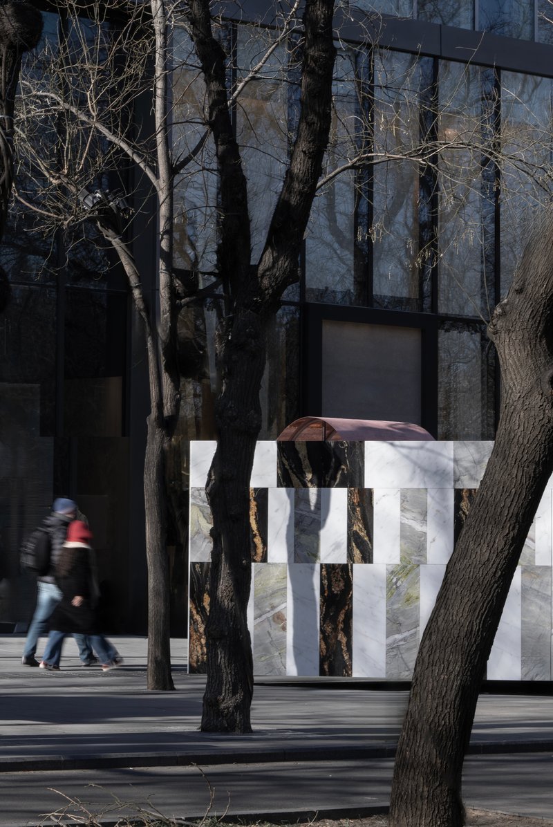 Striped stone wall installation with copper arch element as pedestrians pass beneath bare tree shadows