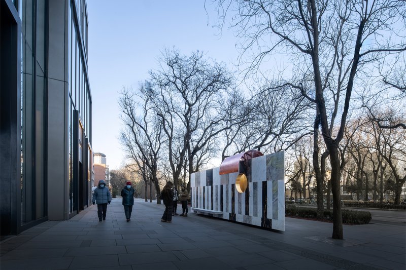 Pedestrians walking along paved plaza with freestanding stone wall and copper tube sculpture at sunset