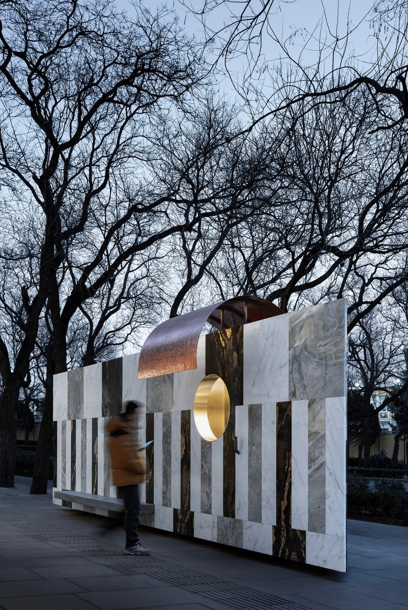 Freestanding wall of vertical stone stripes with copper canopy and pedestrian passing through at twilight