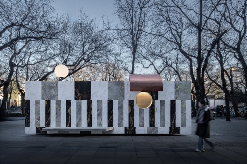 Freestanding pavilion with alternating marble panels and copper canopy in a public plaza with bare winter trees