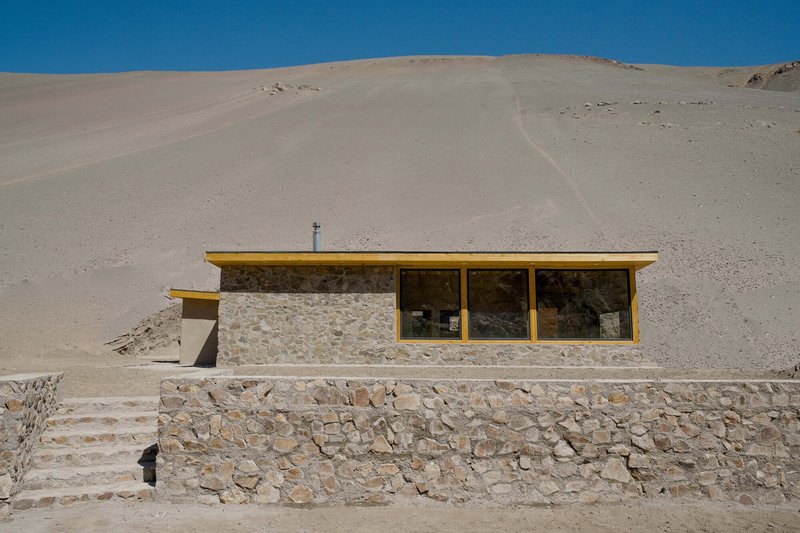 Stone wall structure with yellow-framed ribbon windows set against a barren desert hillside under bright blue sky