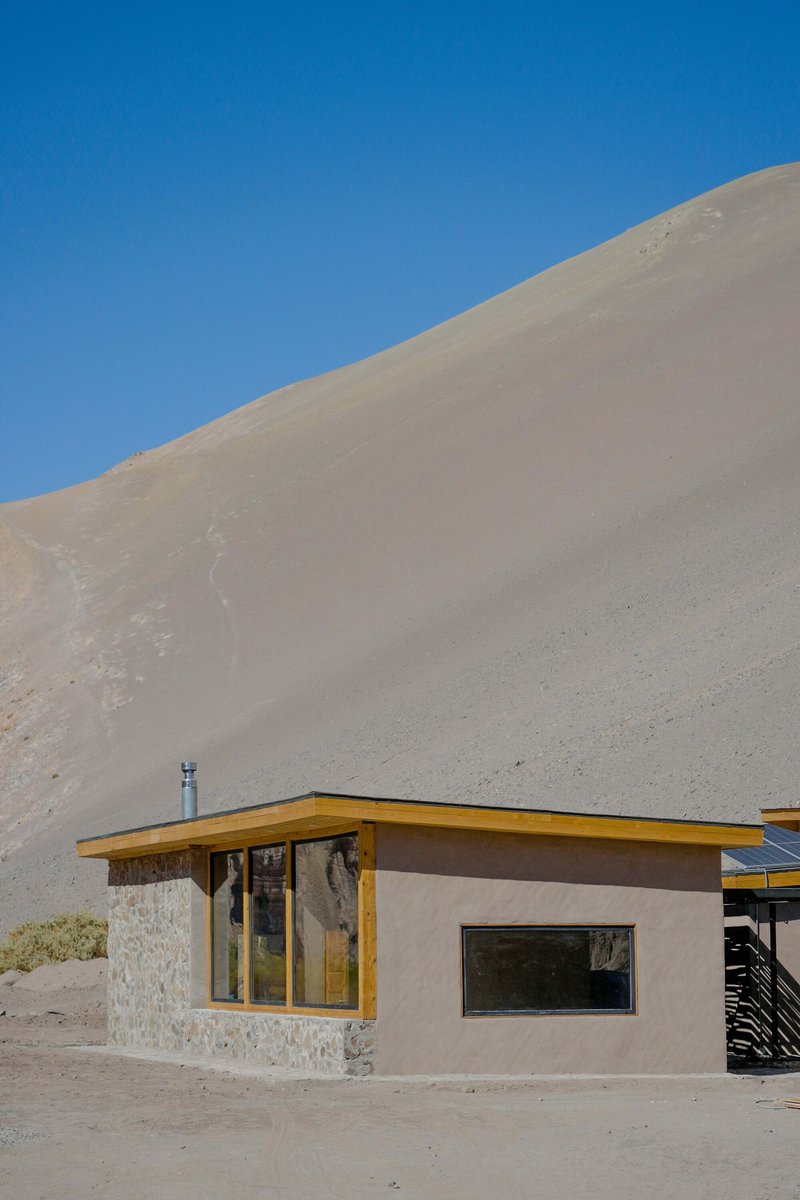 Low pavilion with stone base and timber-framed windows set against a towering sand dune under clear blue sky