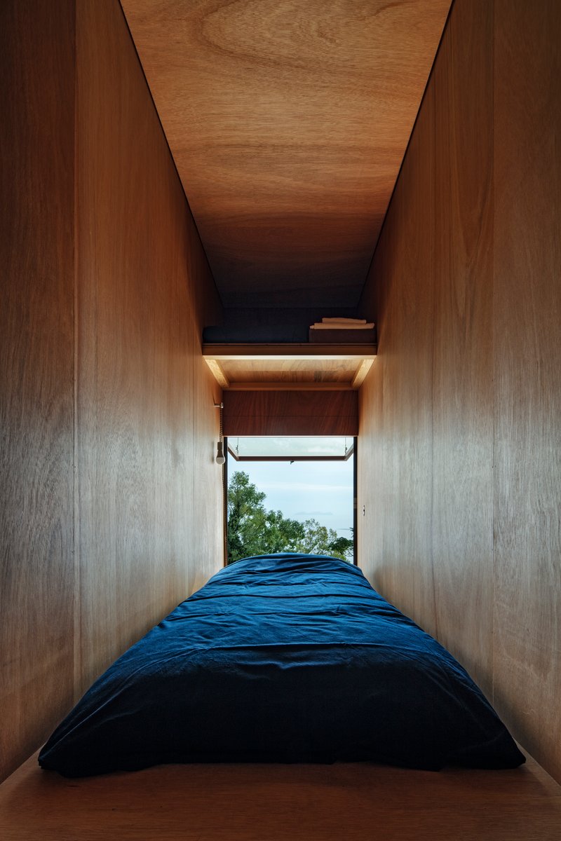 Narrow sleeping nook with plywood walls and ceiling opening to a window with ocean view
