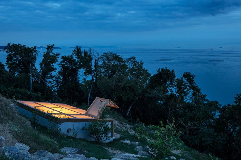 Cantilevered roof illuminated from within overlooking the forested coastal hillside at night