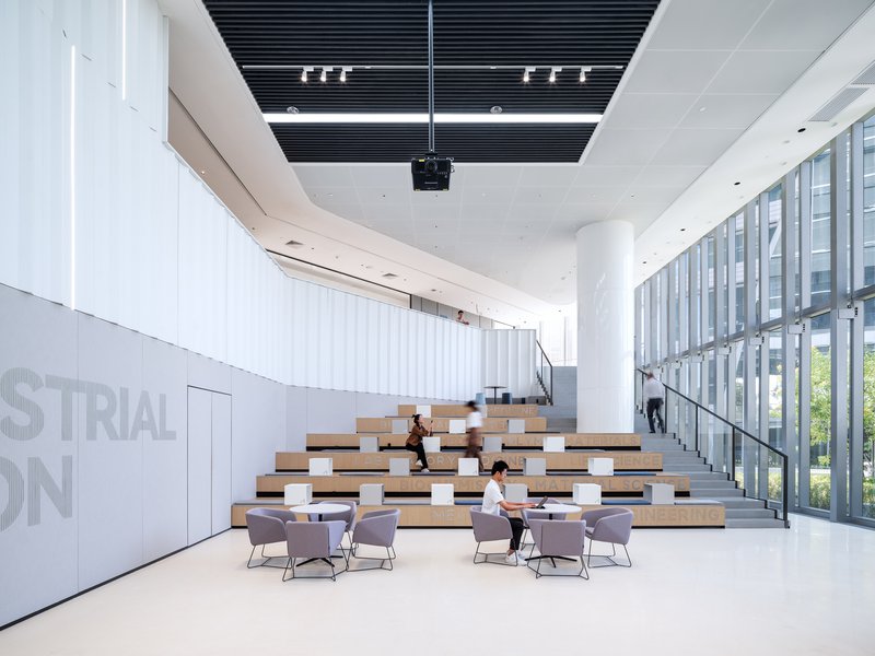 Double-height lobby with tiered wooden seating and people seated in grey chairs below