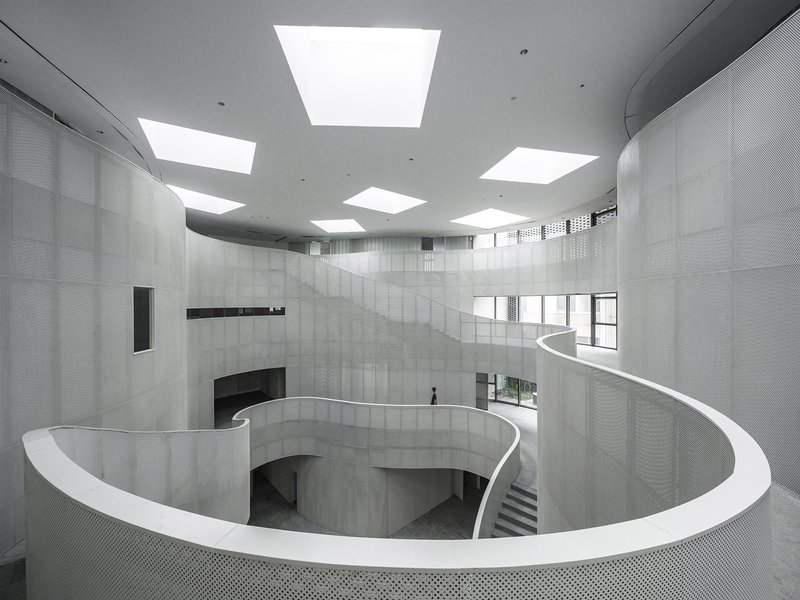 Spiraling white balustrade and concrete walls beneath square skylights in a multi-story atrium with a lone figure