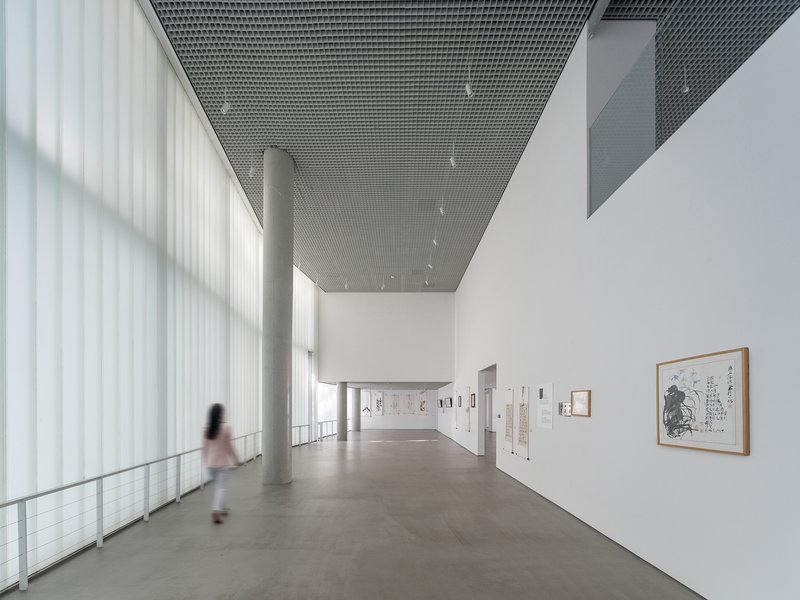 Gallery with perforated metal ceiling, translucent curtain wall and a figure walking past framed artworks