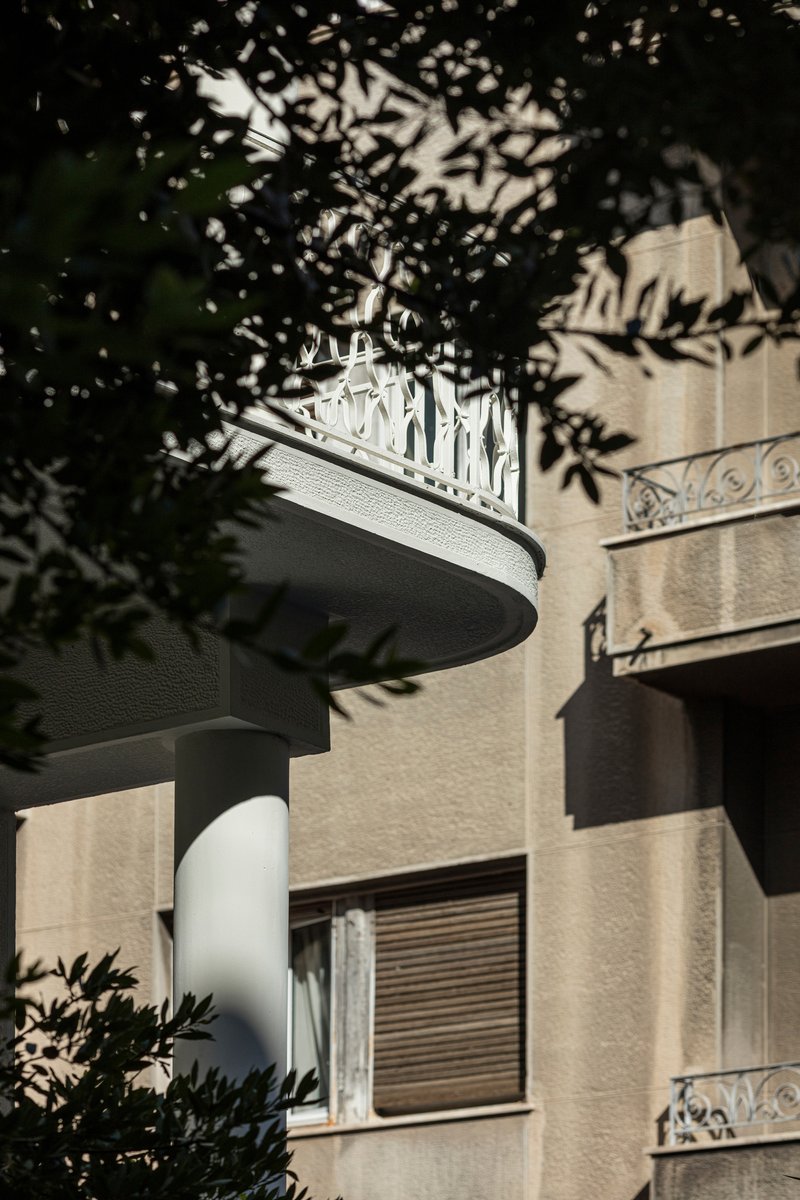 Curved balcony with decorative metal railing visible through dappled tree shadow cast on adjacent beige facade