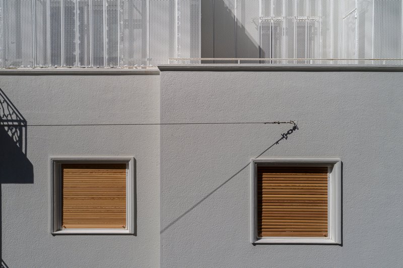Two square windows with timber roller shutters set into pale rendered wall with diagonal shadow line