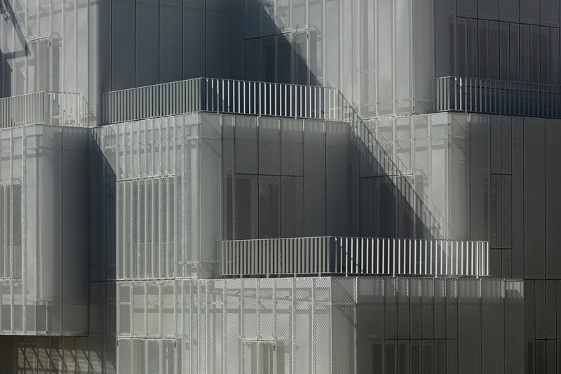 Offset balconies with slatted railings recessed into perforated metal screen facade with shadow patterns across surface