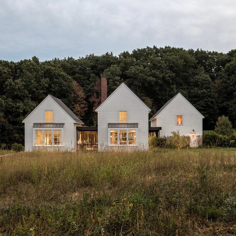 Three white-clad gabled volumes with lit windows across a meadow at dusk