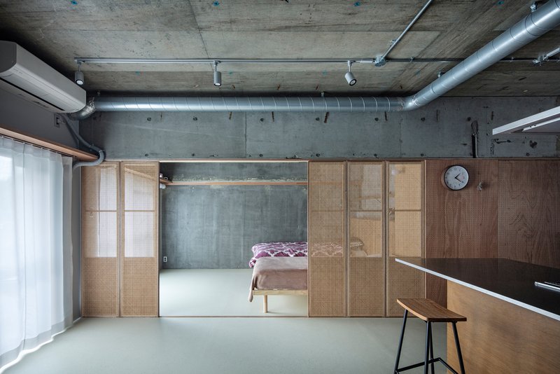Sliding timber and cane panels framing a bedroom alcove beneath an exposed concrete ceiling with metal ducts