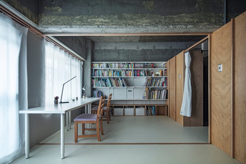 Work area with white desk and open bookshelf against raw concrete walls under natural light from curtained windows