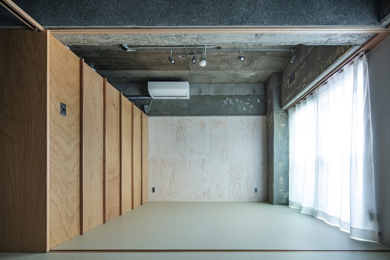 Top-down view of plywood-clad storage wall and grey resin floor lit by diffused daylight through sheer curtains