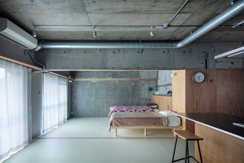 Bedroom with exposed concrete ceiling, metal ductwork, wooden storage wall and pale resin floor