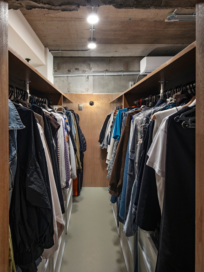 Narrow walk-in closet with hanging garments on both sides under exposed concrete ceiling with track lighting