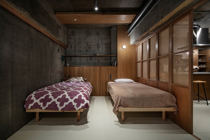 Sleeping area with two platform beds, cane-paneled timber storage wall and exposed concrete ceiling with track lighting