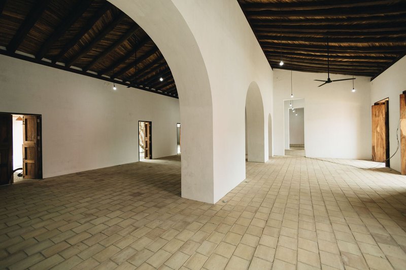 Interior gallery space with arched openings in white walls and woven bamboo ceiling above clay tile floor