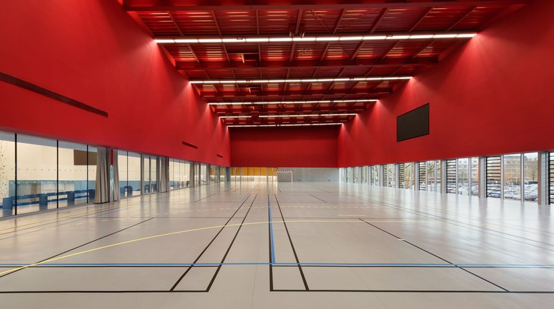 Gymnasium court with red-lit coffered ceiling and glazed wall overlooking adjacent courtyard