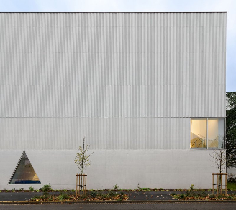 Composite facade with vertical corrugated metal panels above smooth rendered base and triangular corner window at dusk